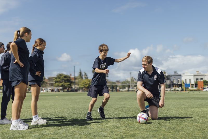 Henry kneels beside a soccer ball while a young student winds up to kick it. Other students are queuing for their turn.