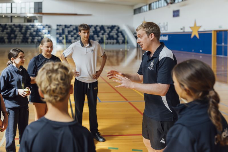 Henry stands in a circle with students inside a gymnasium, holding out his hands in readiness to catch a ball that a student is about to throw to him.