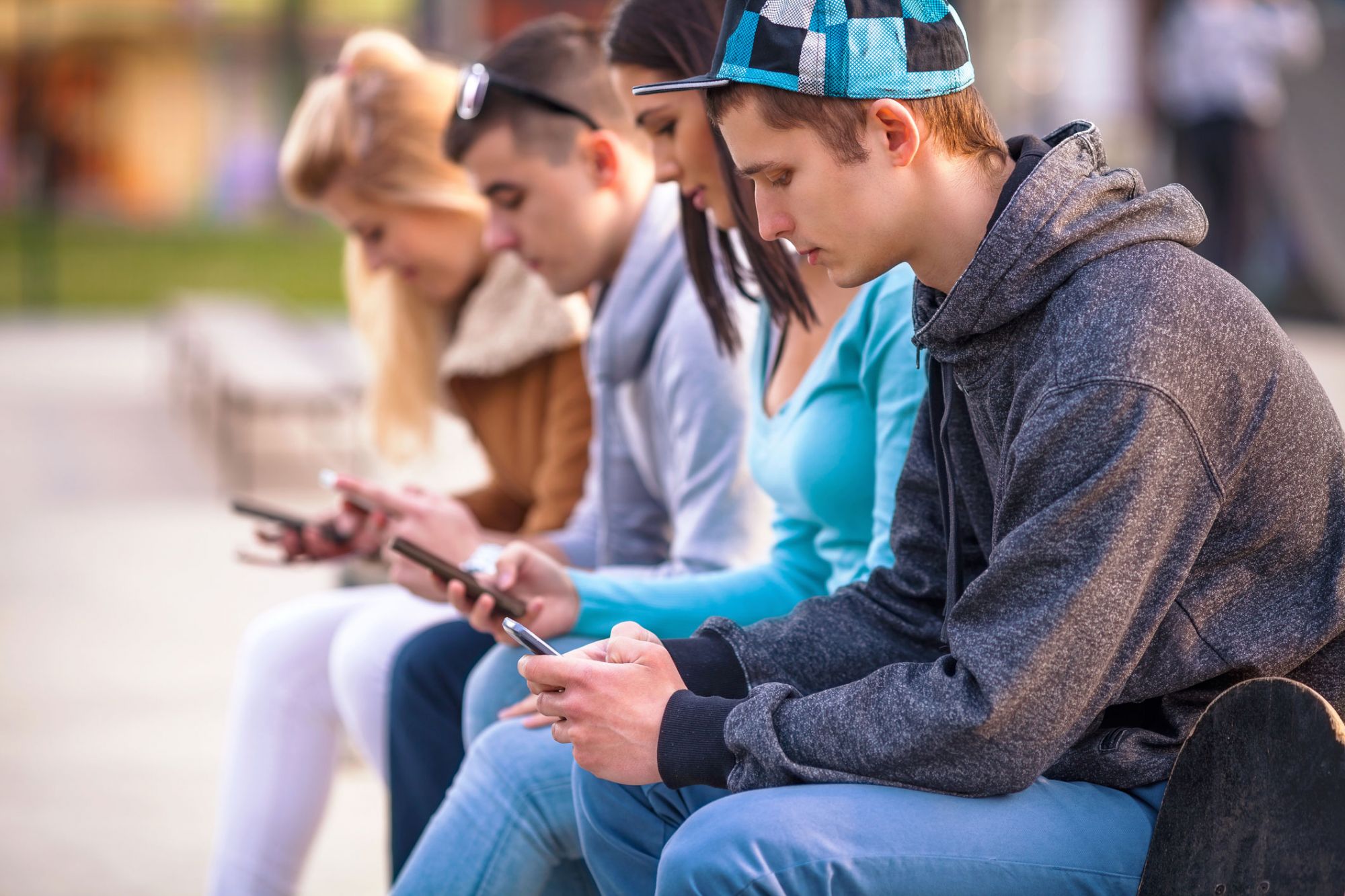 four students sitting on park bench looking at their mobile phones