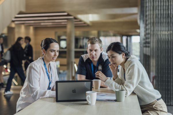 Three young teachers seated at a table look at a laptop screen and plan together.