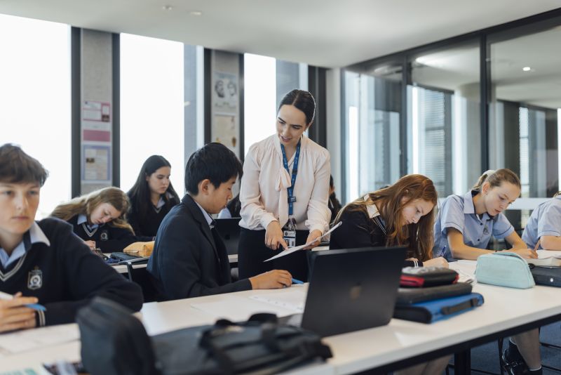 As students work at their desks, Elysia stands beside one student and explains something about the worksheet, pointing something out to him.