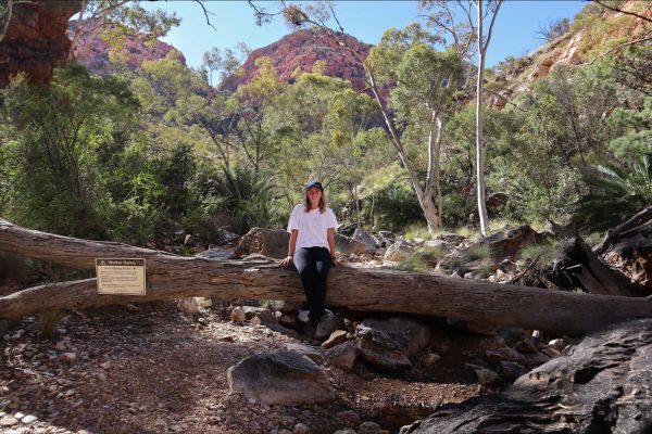 A woman sits on a fallen log across a rocky dry creekbed in the Northern Territory. Behind her is scrubby greenery, and behind that, the peaks of tall red rock hills can be seen against a blue sky.