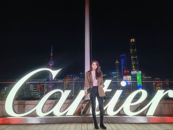 Cara stands in front of large glowing lettering that reads 'Cartier', with a city skyline at night shown through the window behind her.